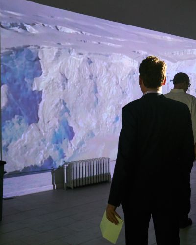 Nearly full-body photograph of a gallery-goer's back as he looks at a large wall with an image of a large glacier on it. He is holding a yellow brochure of sorts in his left hand. A few other non-descript people are also in the frame.