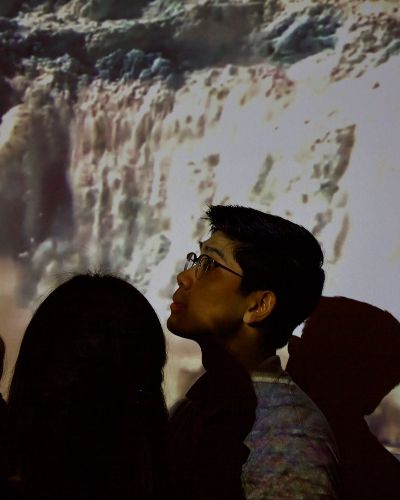 Close-up photograph of the profile of a gallery-goer's head, illuminated by a projection, in the foreground. In the background is a soft, white, projected image of a large glacier calving.