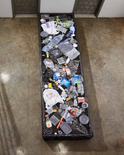 Photograph of top-down view taken from a top a staircase. On the floor below is a ten-by-three-foot small pool, one foot high, filled with water and over a hundred single-use plastic items. The pool has a black lining so the water appears black. 