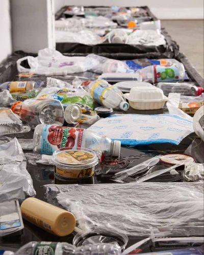 Portrait photograph of hundreds of plastic items floating on a long plane of black water; items get blurry as they recede into a far-off distance