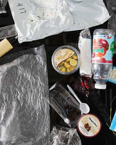 Another tight close-up photograph of about 25 single-use plastic items floating on black water. Most prominently featured is a Tropicana cranberry juice bottle, a lemon dill hummus container, and a Chocolate Chocolate Chip Häagen-Dazs lid.