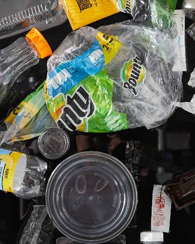 Tight close-up photograph of about 20 single-use plastic items jumbled together. Most prominently featured is a Bounty plastic package wrapping, a Tropicana orange juice bottle, and a circular take-out container.