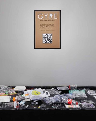 On a gray wall hangs an 18-inch by 24-inch sign that says 'GYRE' followed by some text and a QR code underneath. On the floor, perpendicular to the frame is a one-foot-high black pool with dozens and dozens of single-use plastic items floating on the surface.