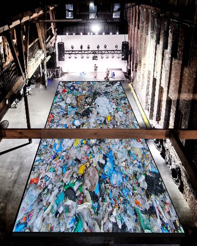 A top-down photograph of a giant, football field-esque gallery space with a giant (2,000 sq feet) rectilinear pool in the middle, packed seemingly thousands of single-use plastic items