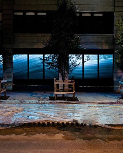 Nighttime photograph showing expansive, 80-foot-long storefront windows with a projection of a geometric rendering of a snowy, bluish, hilly winter landscape that contiguously spans almost the entire width of the storefront. In the foreground, we see a Brooklyn sidewalk fully under construction, with plastic sheeting fully covering the entirety of its surface and a line of &ldquo;CAUTION&rdquo; tape spanning the full length of the frame.