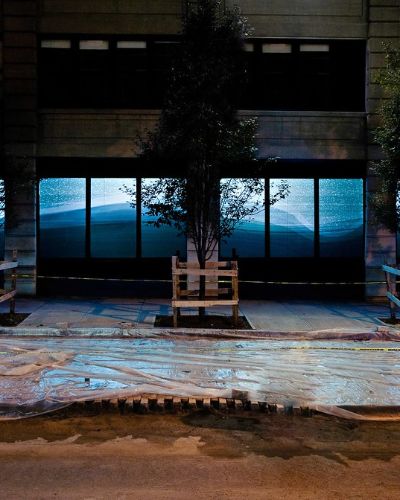 Nighttime photograph showing expansive, 80-foot-long storefront windows with a projection of a geometric rendering of a snowy, bluish, hilly winter landscape in a different geometric configuration than before, which contiguously spans almost the entire width of the storefront. In the foreground, we see a Brooklyn sidewalk fully under construction, with plastic sheeting fully covering the entirety of its surface and a line of &ldquo;CAUTION&rdquo; tape spanning the full length of the frame.