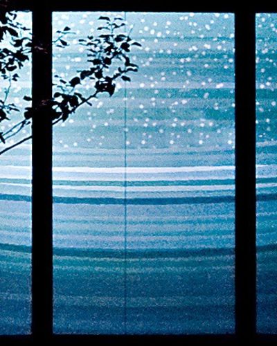 Close-up nighttime photograph of three large storefront windows showing a projection of a concave geometric rendering of a snowy, bluish, and hilly winter landscape. On the left side of the photo, in front of the projection, are branches emanating from a tree without any leaves.