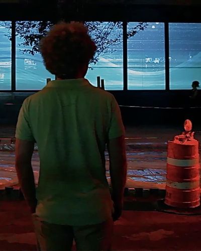 Nighttime photograph of the back of a man, centered in the frame; behind him is an illuminated storefront window on a street with lots of construction (orange work-in-progress construction cone to his right). Projected in the windows is a hilly winter scene, a geometric rendering of layers of ice stacked on top of each other as snow continues to fall on top; tree branches sans leaves are silhouetted in front of the projection.