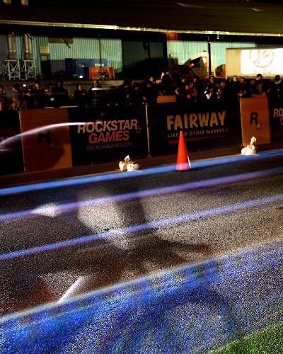 Nighttime, long-exposure photograph showing the &ldquo;light lines&rdquo; of two racing cyclists with a large crowd behind them. Onto the street on which they race is projected a linearly oriented rainbow road of colored lanes.
