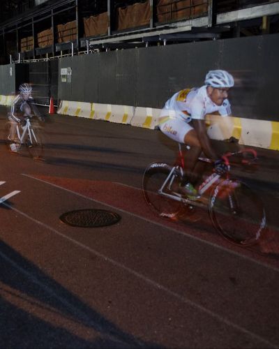 Nighttime photograph of three cyclists, one behind the other, racing on a street course. Underneath the lead cyclist's bike is a thin, illuminated red lane.
