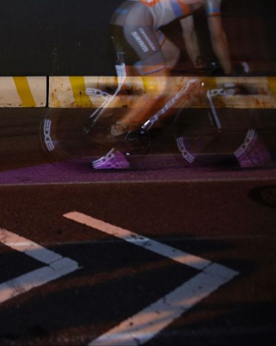 Nighttime, close-up photograph of a street with a thin purple lane projected on it. Riding over the purple lane is a cyclist, whose white cycling shoe is also covered in purple light.