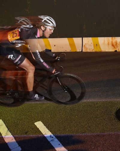 Nighttime photograph of one cyclist, seen from the side, racing on a street course. Underneath their bike is a thin, illuminated yellow lane.
