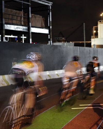 Nighttime, long-exposure photograph showing the 'light lines' of five racing cyclists with a large crowd behind them. Onto the street on which they race is projected a linearly oriented rainbow road of colored lanes