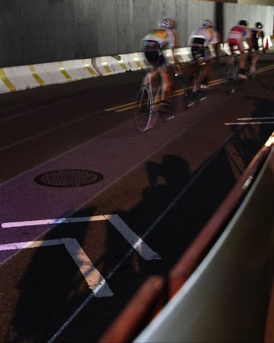 Nighttime photograph of four cyclists, one behind the other, racing on a street course as seen from behind. Underneath the last two cyclists' bikes is a thin, illuminated purple lane.
