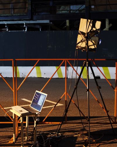Nighttime photograph showing an orange racing barrier/fence that spans the full length of the frame. In front is a chair with an open laptop on a stand connected to a projector on a high tripod. In the background is an empty race course. There aren't any people in the photograph.