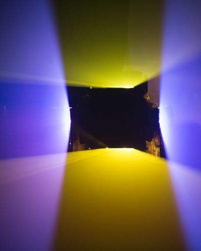 Photograph of a dark room lit by two vertically oriented yellow planes of light and two horizontally oriented purple planes of light. The planes intersect each other to form a black, hollow square shape (where there isn&rsquo;t any light) in the middle of the frame.