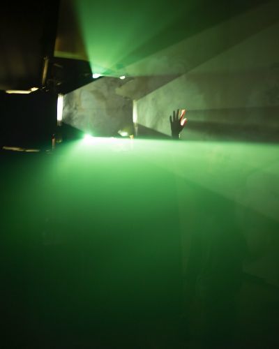 Photograph of a dark room with a strong light source (projector) on the left side of the frame. Emanating from the projector, in a fog-filled room, are very prominent and sharply divided planes of pure green light, perfectly parallel to the floor/ceiling and about 18 inches apart. In the gap stretches a child&rsquo;s hand, which creates its own sharply defined shadows in the opposite direction of the light source.