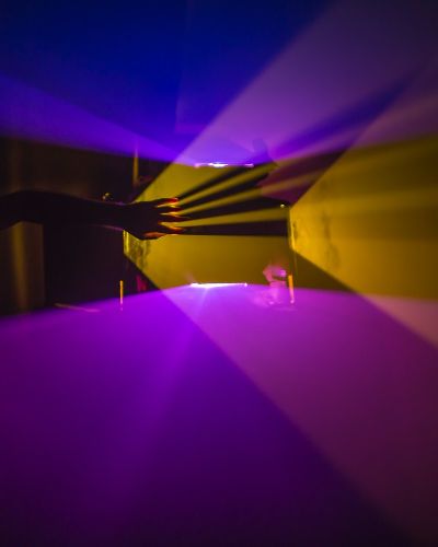 Photograph of a dark room with a strong light source (projector) on the left side of the frame. Emanating from the projector, in a fog-filled room, are very prominent and sharply divided planes of purple and orange light that form V-shapes as they expand outward. In the middle of the frame, a hand reaches up from the shadows and creates its own sharply defined shadows in the opposite direction of the light source.