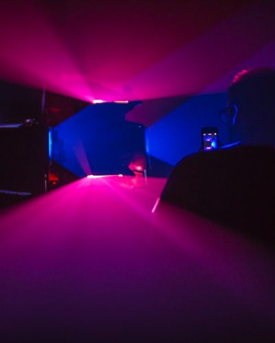 Photograph of a dark room lit by two vertically oriented lavender planes of light and two horizontally oriented yellow planes of light. The planes intersect each other to form a black, hollow square shape (where there isn&rsquo;t any light) in the middle of the frame.