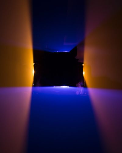 Photograph of a dark room lit by two vertically oriented orange planes of light and two horizontally oriented blue planes of light. The planes intersect each other to form a black, hollow square shape (where there isn&rsquo;t any light) in the middle of the frame.
