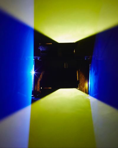 Photograph of a dark, fog-filled room, in the middle of which hover two rectilinearly bound, horizontal planes of yellow light. The planes are roughly 18 inches apart, and in the background is a small crowd of gallery-goers.