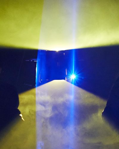 Photograph; planes of yellow light fan out from the top and bottom of the frame; a rectangular black hole in the middle; blue planes of light fan out from the sides. The planes of light reveal fog in the air. Two silhouettes of people with their backs to the camera are probably wondering what the fuck is going on.