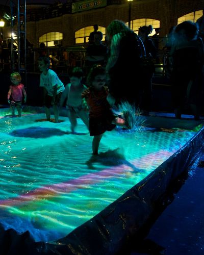Nighttime photograph of a group of children playing in a small pool, the surface of which is being projected onto with a teal green geometric pattern