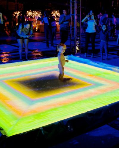 Nighttime photograph of a naked toddler standing in the middle of a small pool, the surface of which is being projected onto with a green, orange, and yellow pattern of concentric rectangles