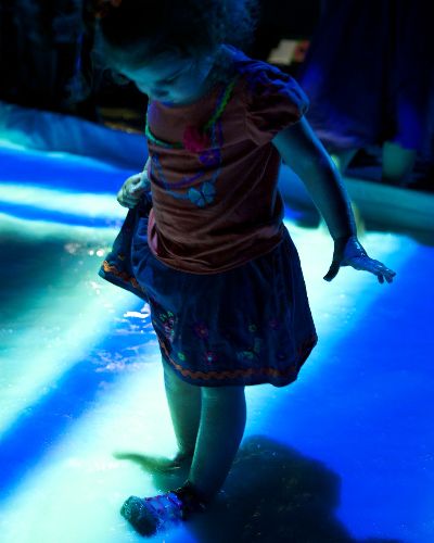 Nighttime photograph of a small girl looking down at her feet which are immersed in a pool of white liquid onto which a blue and pink geometric pattern is projected