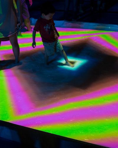 Nighttime photograph of a child in a small pool, the surface of which is being projected onto with a fluorescent pink and green concentric rectilinear pattern with a black diamond at the center, which is where the child is putting their foot