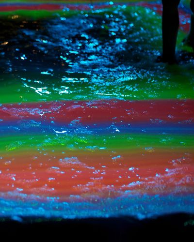 Nighttime close-up photograph of rippling water in a small pool, the surface of which is being projected onto with a geometric rainbow of stacked green, red, blue, and orange rectangles