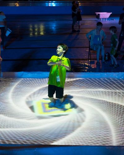 Nighttime photograph of a small pool inside which a small child and an adult play. Projected on the surface is a white spiral of concentric geometric shapes