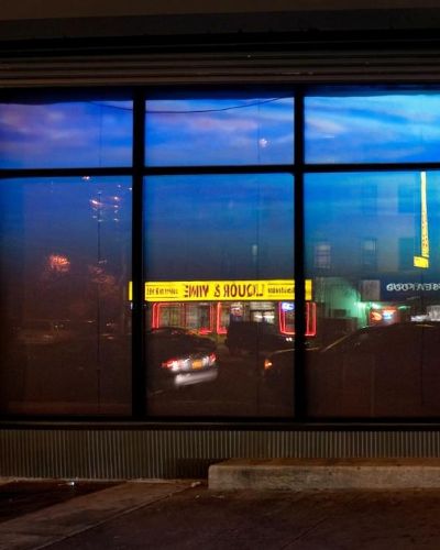 Nighttime photograph of two storefronts, one on each side of the frame, separated by an abandoned-looking door in the middle. In the left storefront is projected a video of the surface of water, from below; reflected in the windows is a bright yellow awning of the liquor store across the street. In the right storefront is projected a violet-ish rendering of clouds as seen from above.