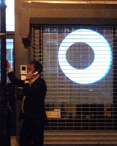 Nighttime photograph of an urban dweller on a cellphone, leaning against a lamppost on the far right side of the frame. Occupying the rest of the frame are two giant cartoon-like eyes that are projected into the gated storefront behind him. The eyes are eyeing him; he has his back to them, seemingly oblivious to their presence.