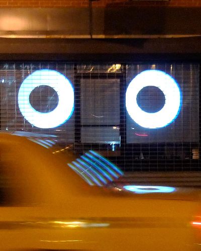 Nighttime photograph of two large, cartoonish eyes projected into a storefront with a metal security gate in the background. In the foreground, a NYC yellow cab whizzes by.