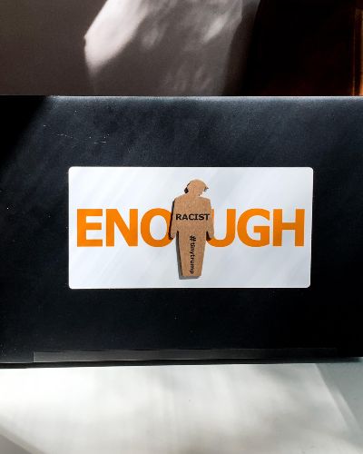 Photograph of a tiny trump ENOUGH sticker on a laptop. The tiny trump stuck in the middle bears the slogan Racist, so the combination reads ENOUGH Racist