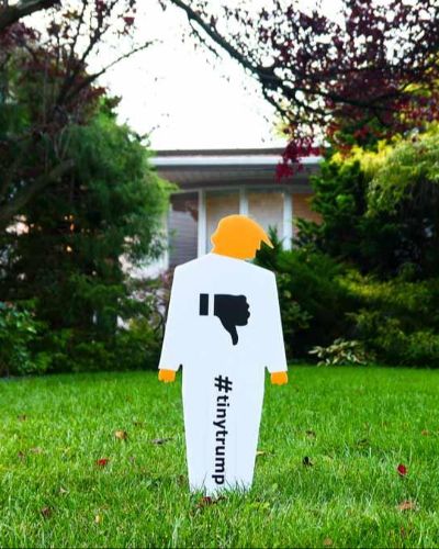 Two-foot-tall lawn sign in the shape of a tiny trump--a cardboard cutout caricature of trump--with a thumbs down sign on the chest. The lawn sign is made out of plastic, is white with orange head and hands. It is mounted on a lawn in front of a suburban house which is in the background.