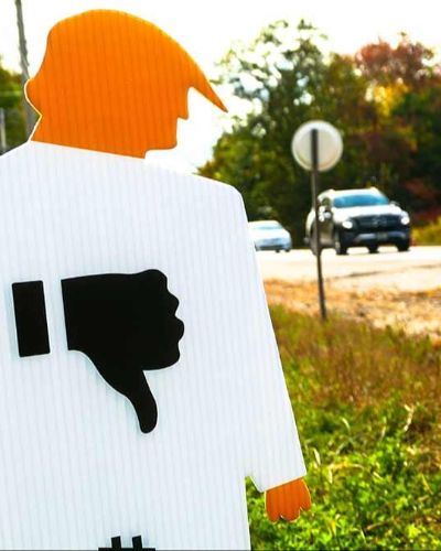 Close up photograph of a tiny trump lawn sign shown on the side of a road. A blurred SUV on a road is in motion in the background.