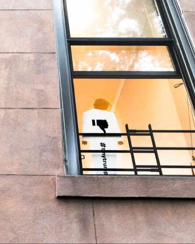 Photograph of a tiny trump lawn shown in an apartment window. The perspective of the photograph is sharply upward, with the tiny trump occuping the lower half of a brownstone apartment window.
