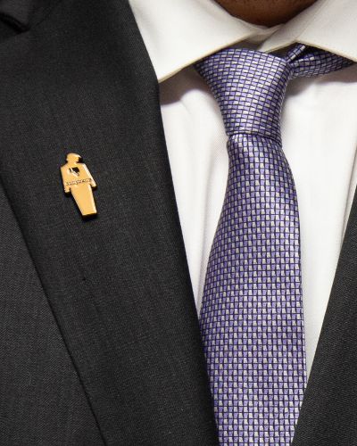 Photograph of a brown tiny trump enamel pin with a thumbs down sign on the left lapel of a man's suit blazer