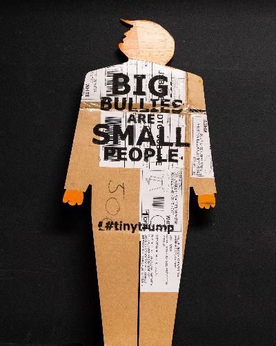 Two-foot-tall caricature cardboard cut out of trump with clown like hands and swooshy hair. Head and hands are painted bright orange and the slogan Big Bullies Are Small People is stamped in black ink across the chest. The cardboard is clearly used--it has shipping labels on it. Gray photo studio background.