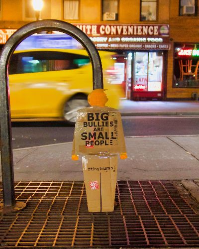 A two-foot-tall cardboard cutout tiny trump protest sign (in the image of a full-bodied trump) leaning against a bike rack in the street in Brooklyn, New York. It is nightime and a yellow cab is blurred in the background as it whizes by.