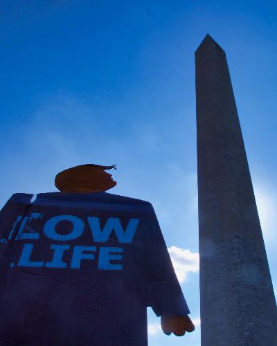 A two-foot-tall cardboard cutout tiny trump protest sign with the slogan Low Life looking up at the Washington Monument in Washington, D.C. Photograph angle is pointed up sharply so only the tiny trump, monument, and sky are visible