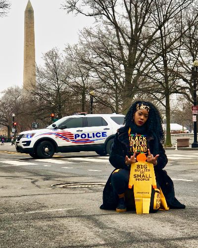 Photograph of a Black female posing with a tiny trump with the slogan Big Bullies Are Small People; she is giving the tiny trump the middle finger with both hands. In the background a police car passes by and further in the background is the Washington Monument.