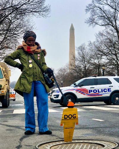 Photograph of a Black female 'walking' a tiny trump that says Dog with a dog collar. The Washington Monument is in the background.