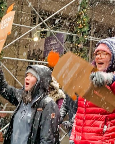 Photograph from the side of two students marching through a very windy and snowy New York City street, each holding a tiny trump