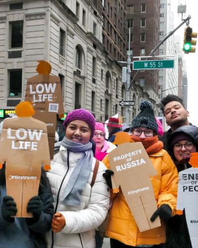 Group photograph 8 people, including the artist, holding up tiny trumps during the protest march, in a NYC intersection. In the background the street sign denoting 55th street is visible.