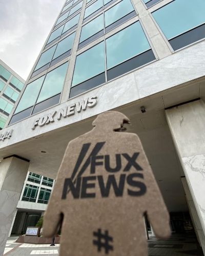 a photograph of a tiny trump with the slogan 'Fux News' held in front of Fox News headquarters in NYC