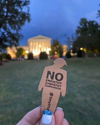 a photograph of someone holding a tiny trump with the slogan 'No Empathy, No Sympathy, No Compassion' in front of the White House