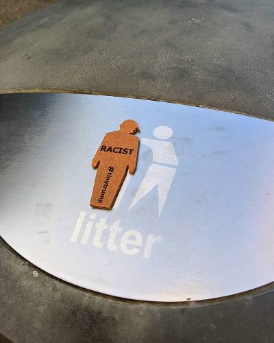 a photograph of a tiny trump with the slogan 'Racist' stuck to a garbage can that's labeled 'Litter'